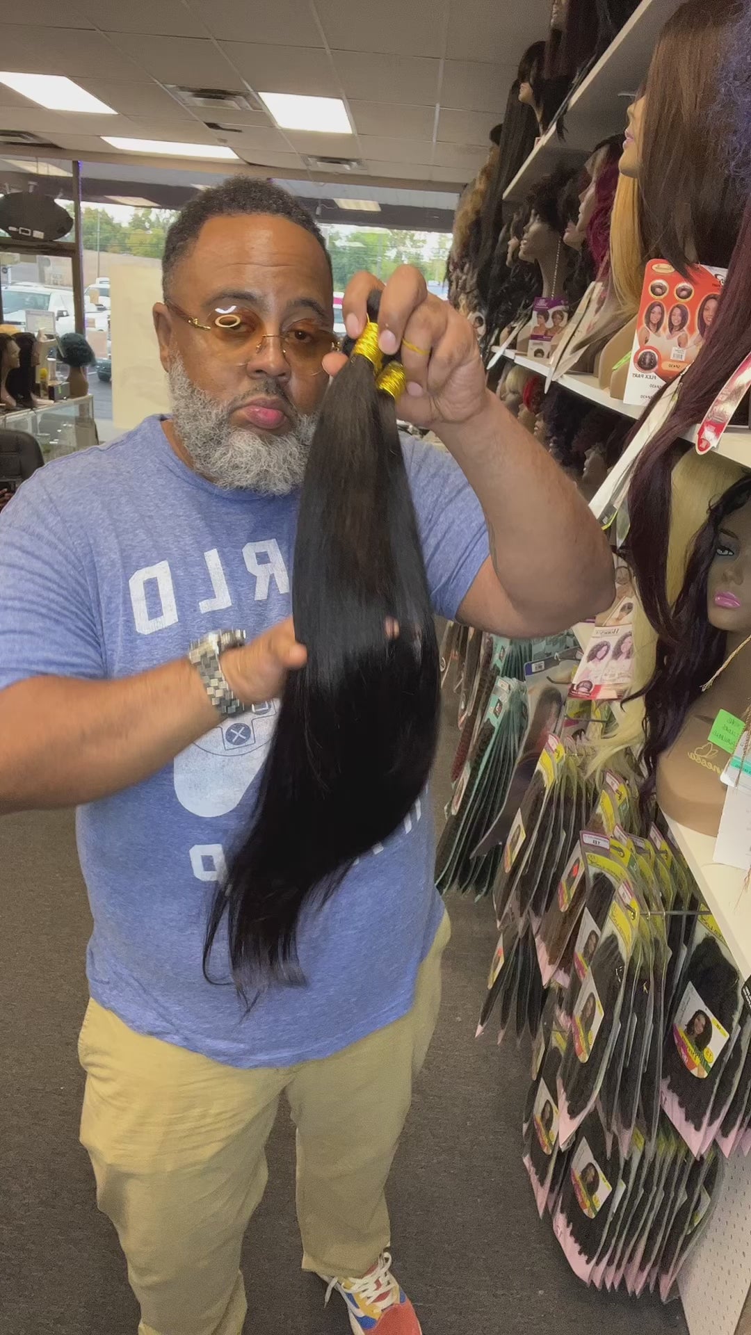 Inside hair Video of handsome man holding 26”28”30” bone straight virgin Brazilian hair bundle deal inside hair store with multiple wigs and beauty products in background 