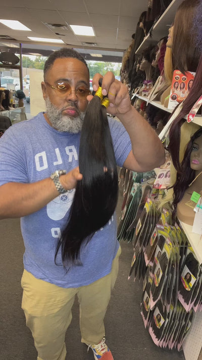 Inside hair Video of handsome man holding 26”28”30” bone straight virgin Brazilian hair bundle deal inside hair store with multiple wigs and beauty products in background 